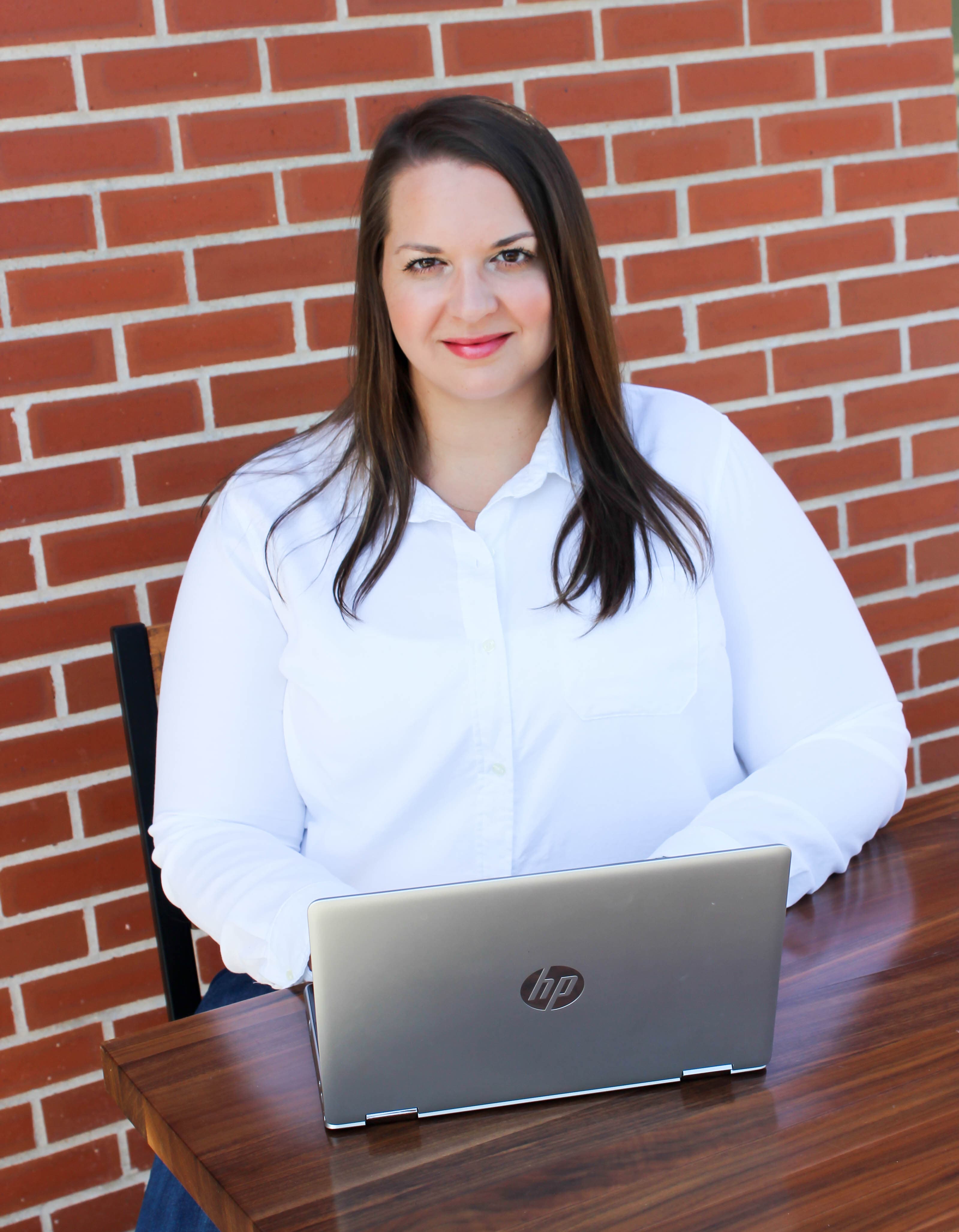 Woman in white shirt using a laptop at a wooden table against a brick wall.
